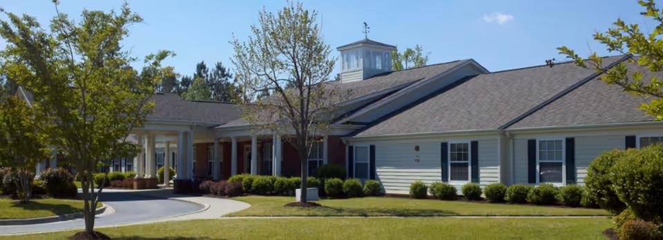 Exterior view of a single-story senior living facility building with a covered entrance, surrounded by trees, shrubs, and a well-maintained lawn under a clear blue sky.
