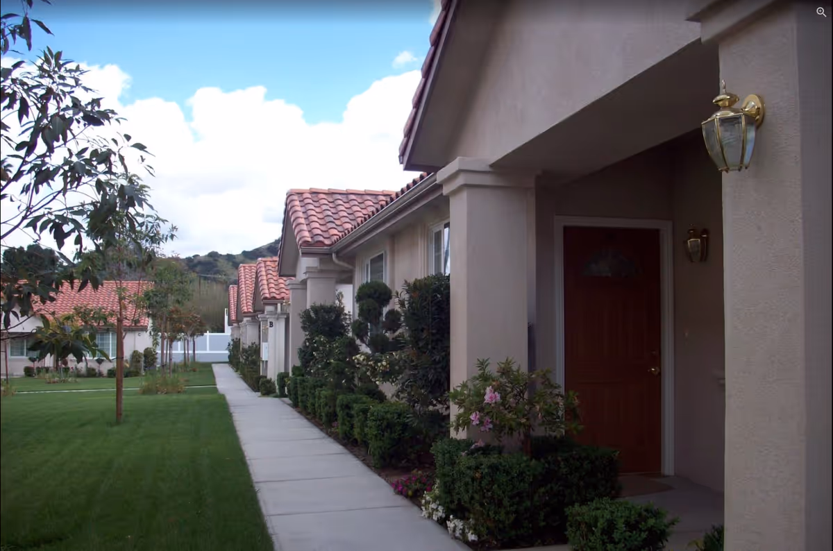 A row of single-story residential units with beige stucco walls and red tile roofs, lined along a concrete walkway. The walkway is bordered by neatly trimmed bushes and small trees, with a well-maintained green lawn on the left side. The sky is partly cloudy with blue patches visible.