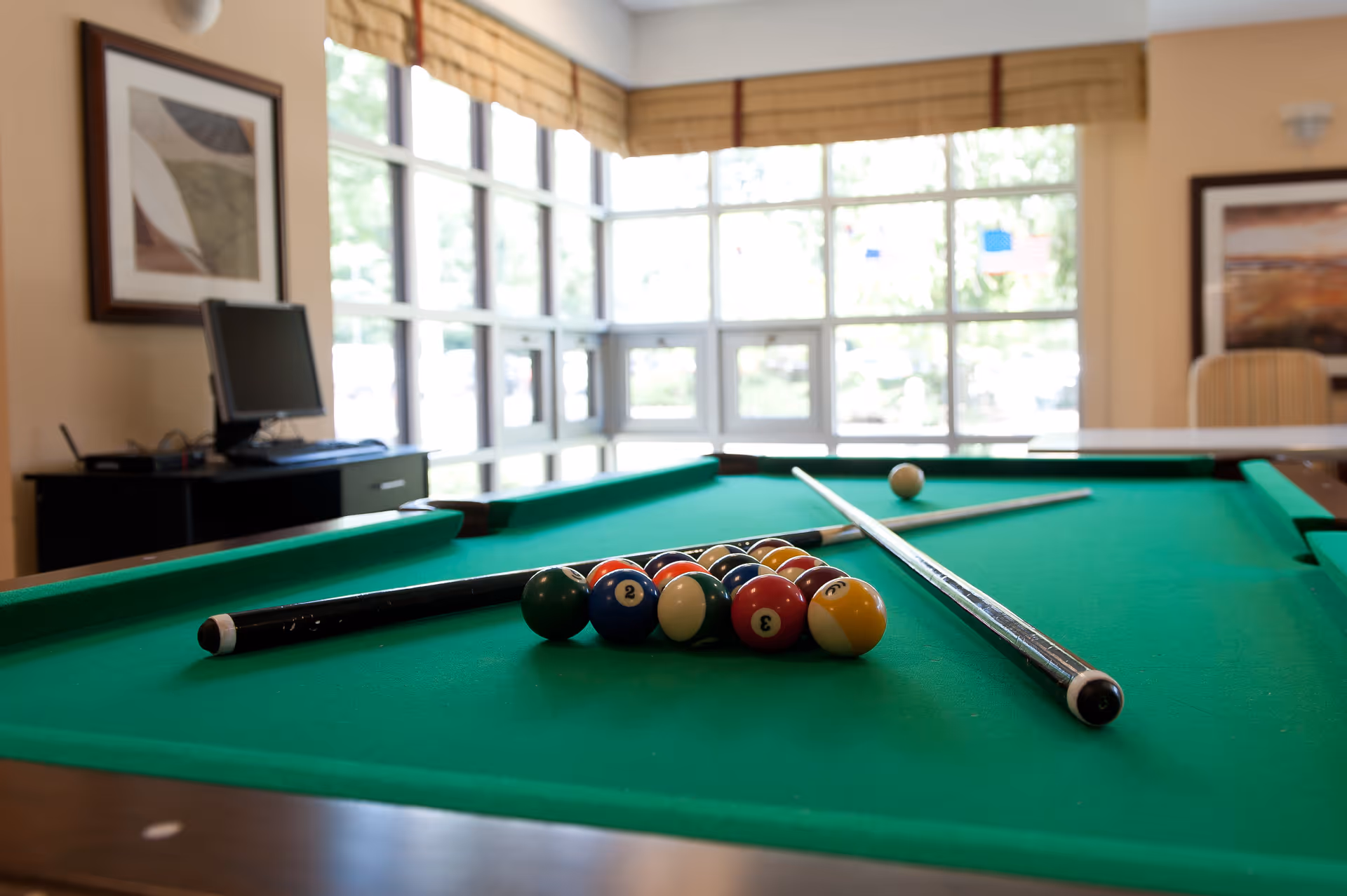 A pool table with racked billiard balls and cues in a bright recreation room with large windows.