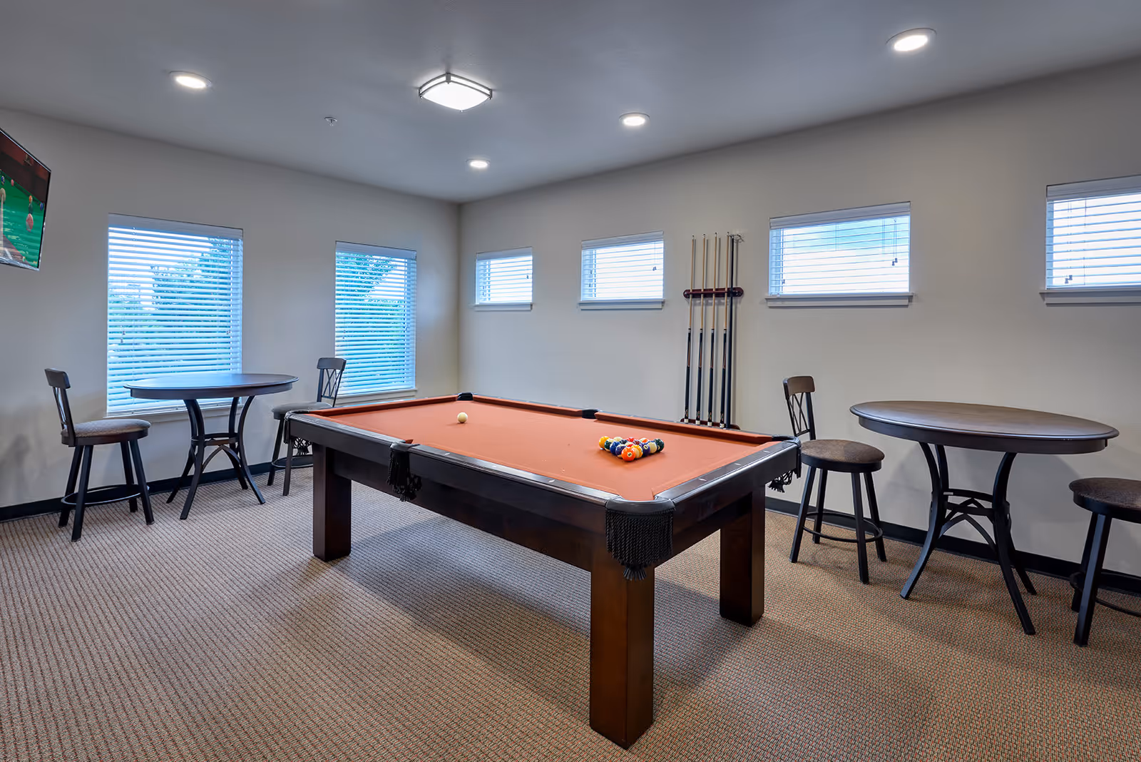A well-lit game room with a pool table in the center, featuring a red felt surface with pool balls arranged for a game. The room has multiple windows with blinds, two round tables each accompanied by two chairs, and a wall-mounted television showing a sports game. Pool cues are neatly arranged on a rack on the wall.