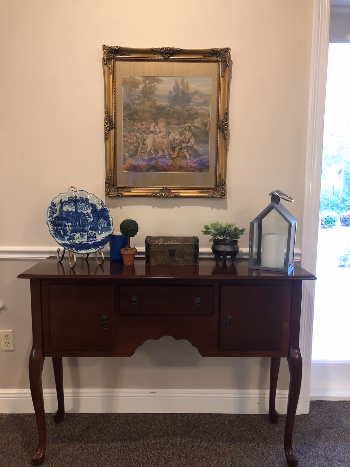 A wooden side table with curved legs against a beige wall. On the table are decorative items including a blue and white plate on a stand, a small potted topiary, a small chest, a potted succulent, and a large lantern with a candle inside. Above the table hangs a framed painting depicting children and a lamb in a pastoral setting. To the right, there is a glass door showing some greenery outside.