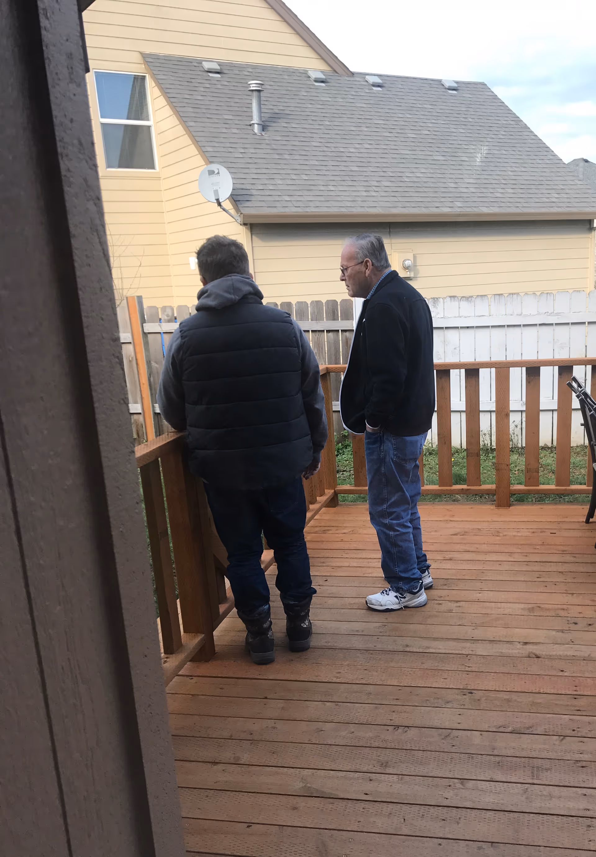 Two men standing on a wooden deck outside, facing away from the camera. One man is wearing a black puffer vest and dark pants, while the other is wearing a black jacket, blue jeans, and white sneakers. In the background, there is a beige house with a gray roof, a satellite dish, and a white wooden fence.