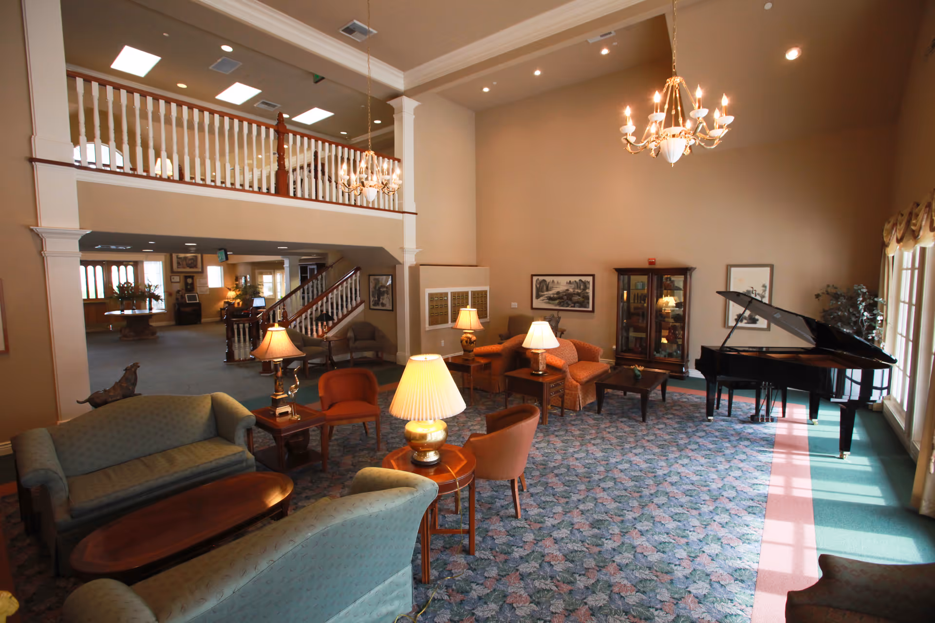 A spacious living room area in a senior living facility with multiple seating arrangements including sofas and armchairs, several table lamps, a grand piano near large windows with curtains, a chandelier hanging from the ceiling, and a staircase leading to an upper level with a railing.