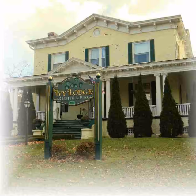 Front exterior of a yellow two-story house with a porch and a green sign reading "Ivy Lodge Assisted Living" on the lawn.