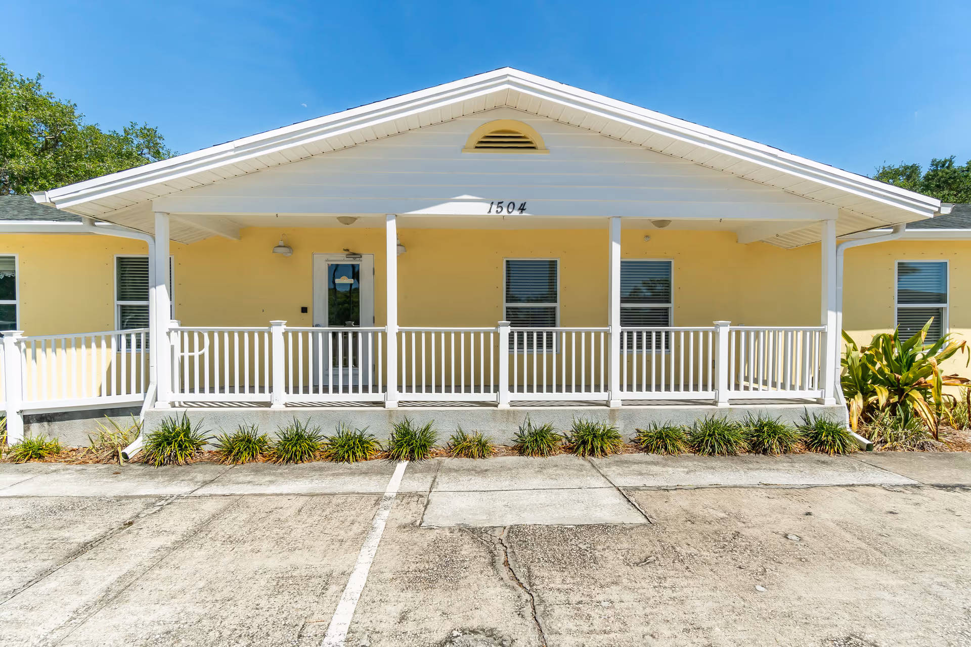 Front exterior view of a single-story yellow building with a white porch railing and a covered entrance. The building has three windows and a door, with the number 1504 displayed above the porch. There are small plants and shrubs along the front, and a concrete parking area in front of the building under a clear blue sky.