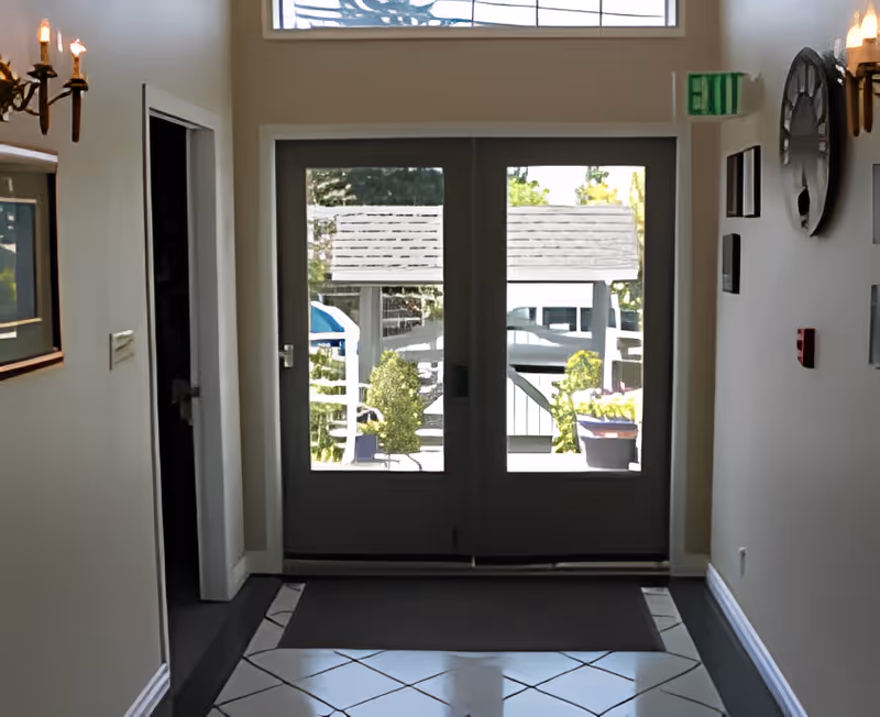 Interior hallway with white walls and tiled floor leading to double glass doors that open to an outdoor area with greenery and a wooden structure. Wall sconces with candles and framed pictures decorate the walls.