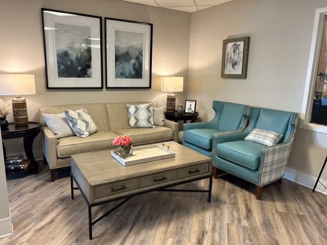 A cozy lounge with a beige sofa, two teal armchairs, a wooden coffee table, lamps, and framed wall art.
