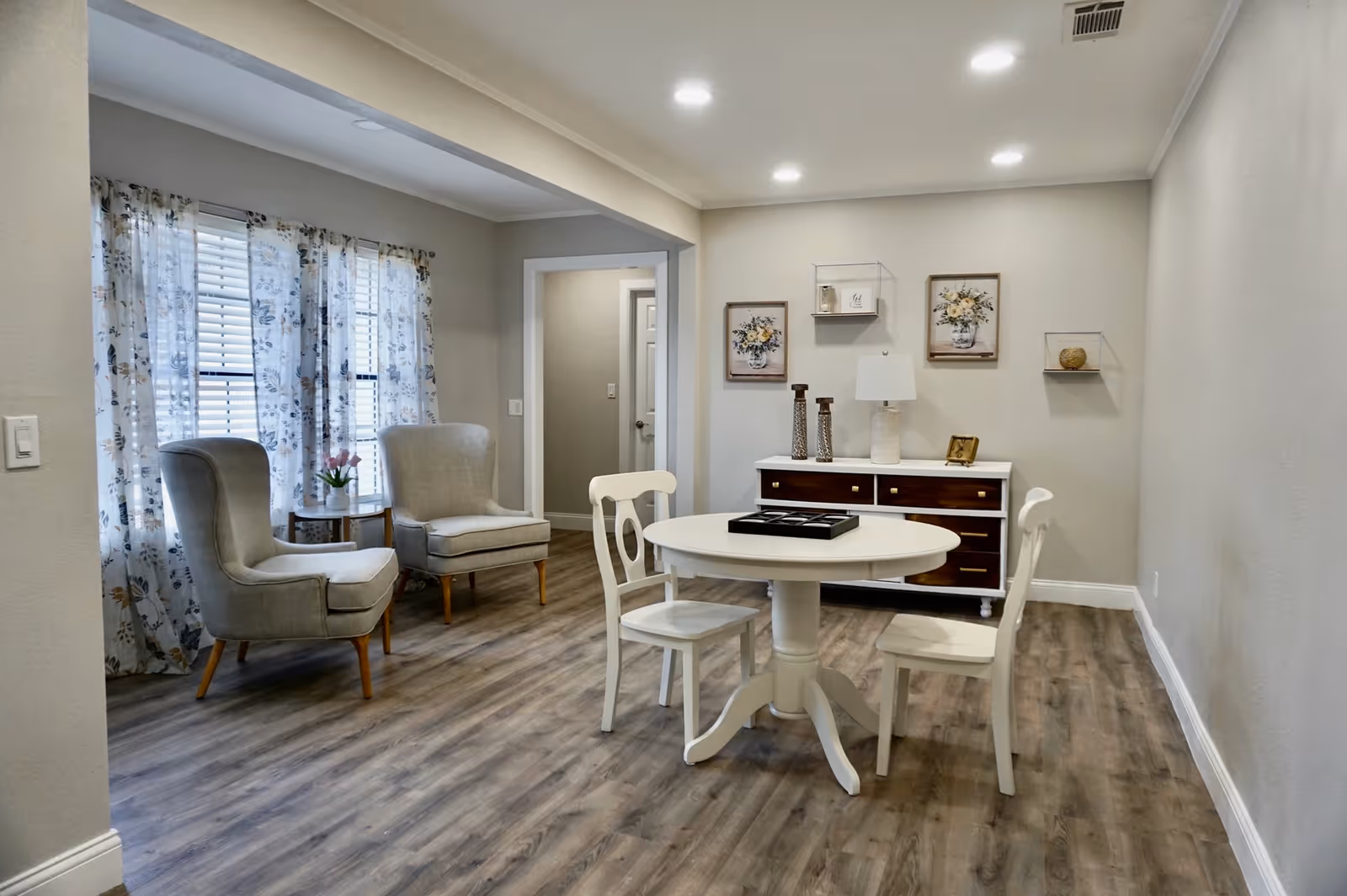 A cozy interior room with two beige armchairs near windows with floral curtains, a small round white table with two white chairs, and a wooden sideboard against the wall decorated with framed floral artwork, a lamp, and decorative items. The room has wood flooring and recessed ceiling lights.