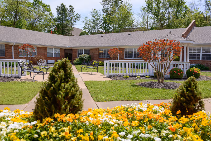 Outdoor courtyard area of a senior living facility with green grass, colorful yellow and white flowers in the foreground, small trees and bushes, benches along a paved walkway, and a single-story brick building with multiple windows in the background under a partly cloudy sky.