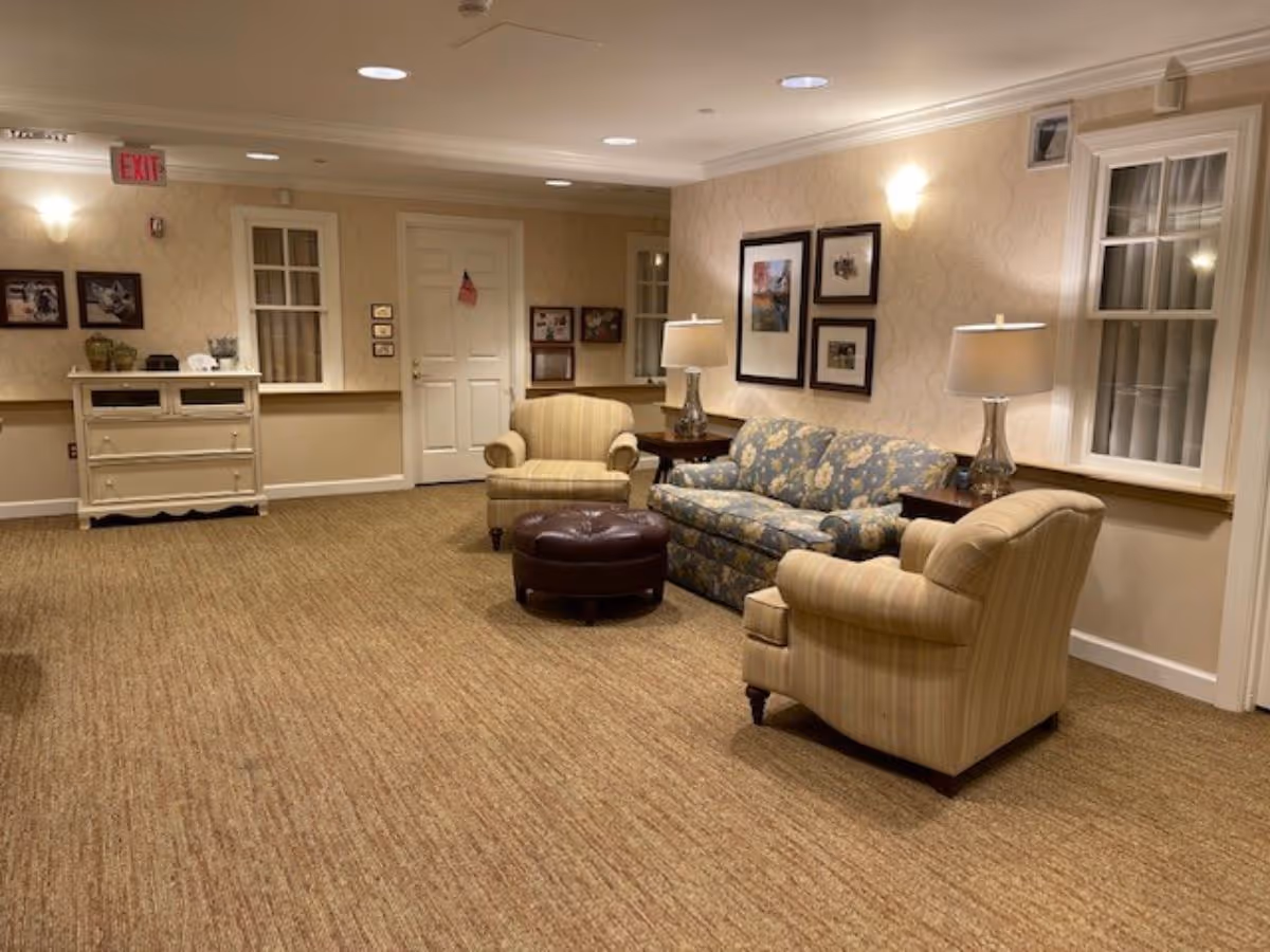 A cozy living room area with beige carpet, two striped armchairs, a floral patterned sofa, a round brown ottoman, two side tables with lamps, framed pictures on the wall, and a white door with an American flag decoration. The room has soft lighting and beige wallpaper.
