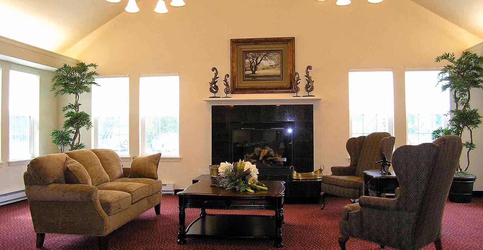 A cozy living room with a brown sofa, two patterned armchairs, a dark wooden coffee table with a floral arrangement, a black fireplace with decorative items on the mantel, and large windows letting in natural light. Two tall potted plants are placed on either side of the room.