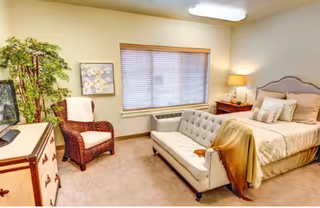 A cozy bedroom featuring a bed with multiple pillows and a beige headboard, a tufted sofa at the foot of the bed with a blanket draped over one arm, a wicker armchair with a white cushion, a wooden dresser with a TV on top, a potted plant, a framed floral artwork on the wall, a window with blinds, and a bedside table with a lamp.