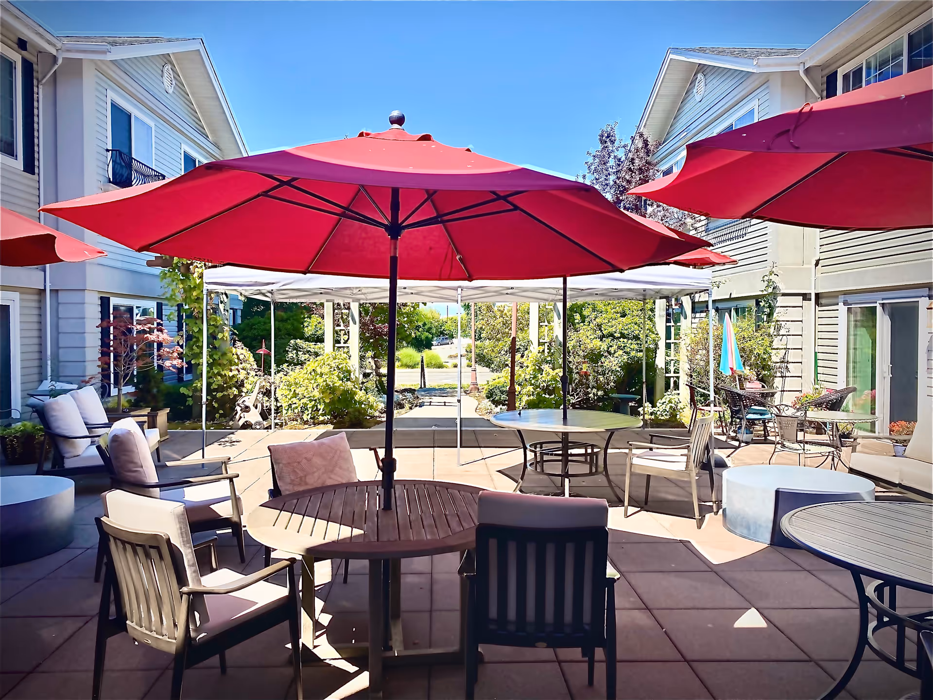 Outdoor patio area at Edmonds Landing by Cogir with multiple round tables and chairs arranged under large red umbrellas. The patio is surrounded by two-story buildings with windows and doors, and there are plants and greenery in the background under a clear blue sky.