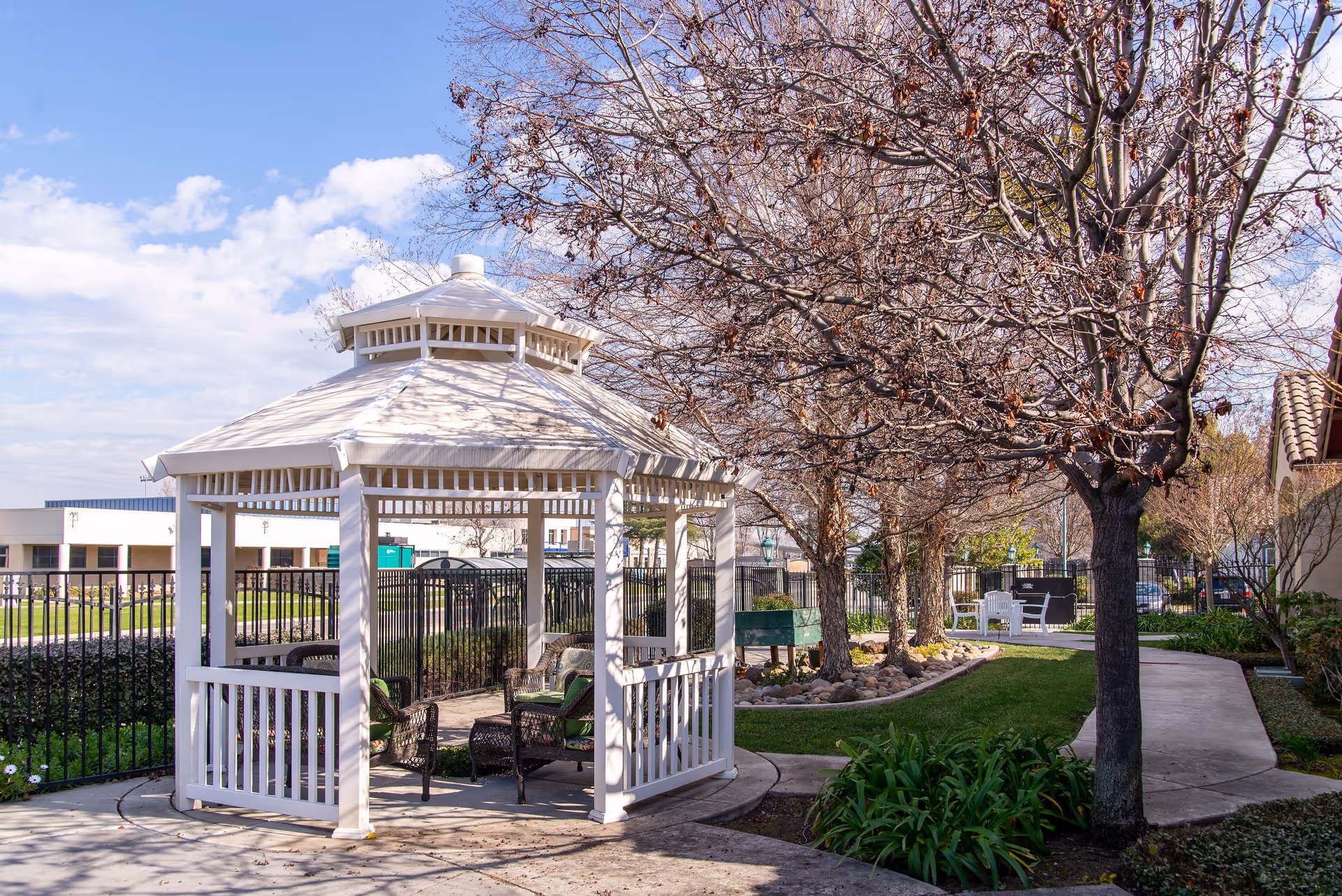 A white gazebo with outdoor seating is situated in a landscaped garden area with leafless trees and a paved walkway. The sky is partly cloudy and the scene appears to be in a senior living facility outdoor space.