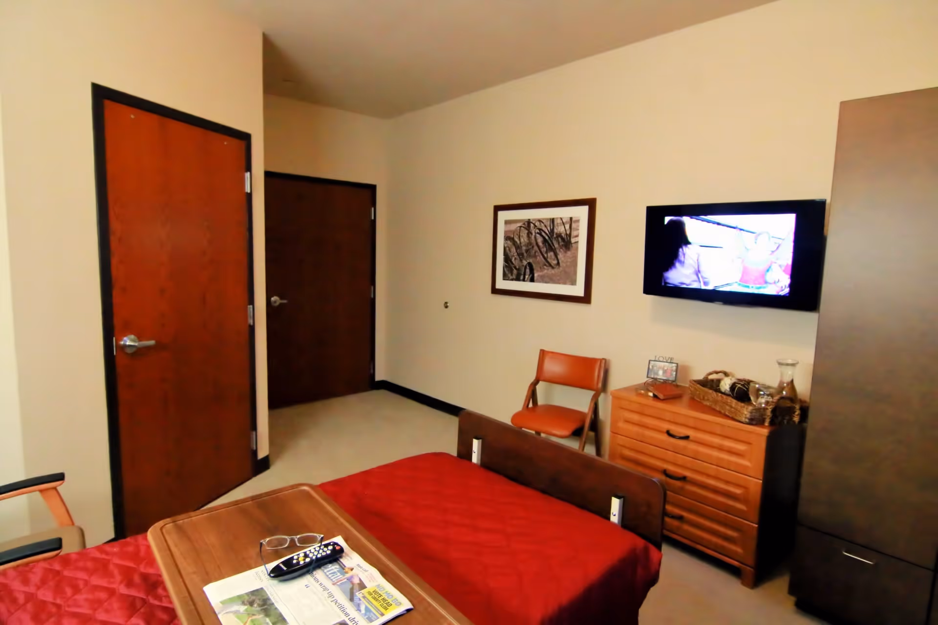 A bedroom in an assisted living center featuring a bed with a red quilt, a wooden tray with reading glasses, a remote control, and a newspaper on it. The room has two wooden doors, a wooden dresser with a basket and decorative items, a wall-mounted TV, a framed picture on the wall, and an orange chair.