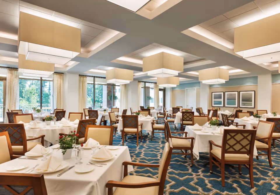 Bright, elegantly furnished dining room with tables set in white linens, wooden chairs, large square pendant lights, and windows overlooking greenery.