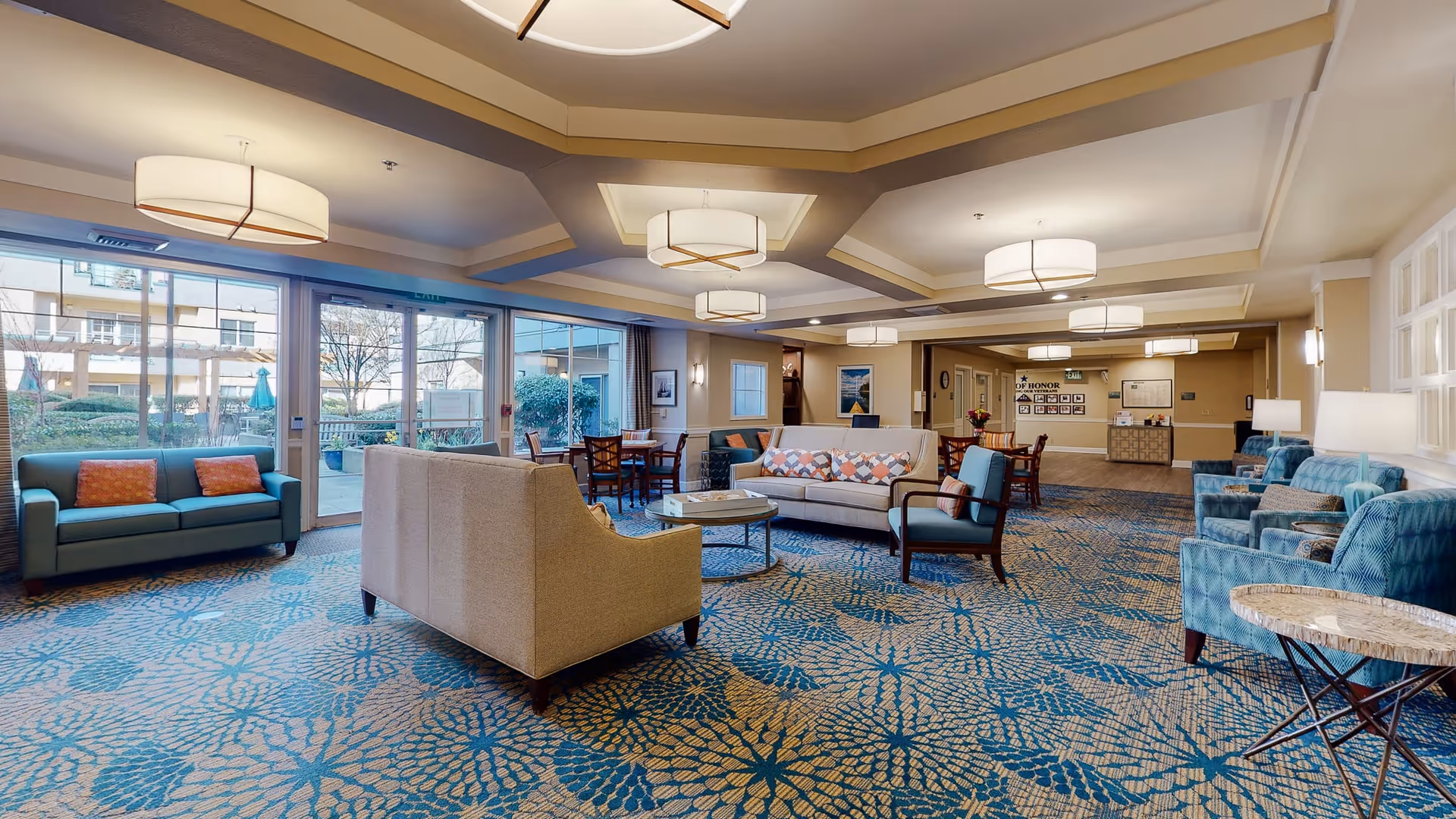 A spacious and well-lit assisted living facility common area with multiple seating arrangements including sofas, armchairs, and small tables. Large windows and glass doors provide a view of an outdoor patio area with greenery. The ceiling features modern light fixtures and the carpet has a blue and beige floral pattern.