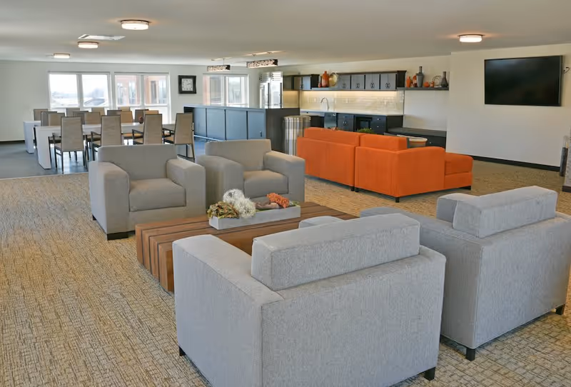 A spacious senior living common area featuring multiple seating arrangements including gray armchairs and orange sofas. There is a wooden coffee table with decorative items, a dining area with tables and chairs near large windows, and a kitchenette with cabinets and a countertop. A flat-screen TV is mounted on the wall.