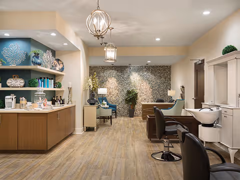 A well-lit interior space featuring a reception or lounge area with wooden flooring. On the left, there is a counter with coffee and beverage supplies, shelves with decorative items and books. In the center and back, there are comfortable chairs and a small table against a textured stone accent wall with plants and lamps. On the right, there are two black salon chairs and a white sink, suggesting a hair salon or grooming area within the facility.