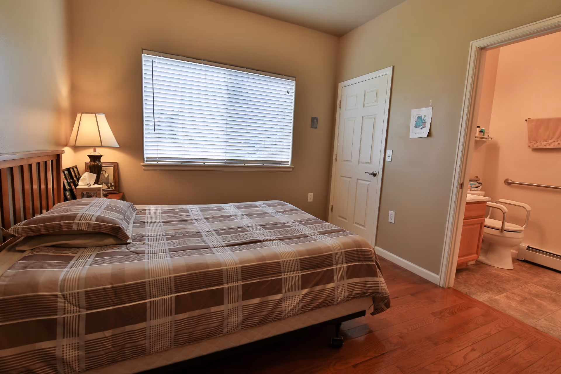 A bedroom with a wooden bed frame and a plaid bedspread. There is a bedside table with a lamp and framed photos. A window with closed blinds is on the wall behind the bed. To the right, there is a door leading to a bathroom with a toilet, grab bars, a towel hanging on the wall, and a sink with toiletries.