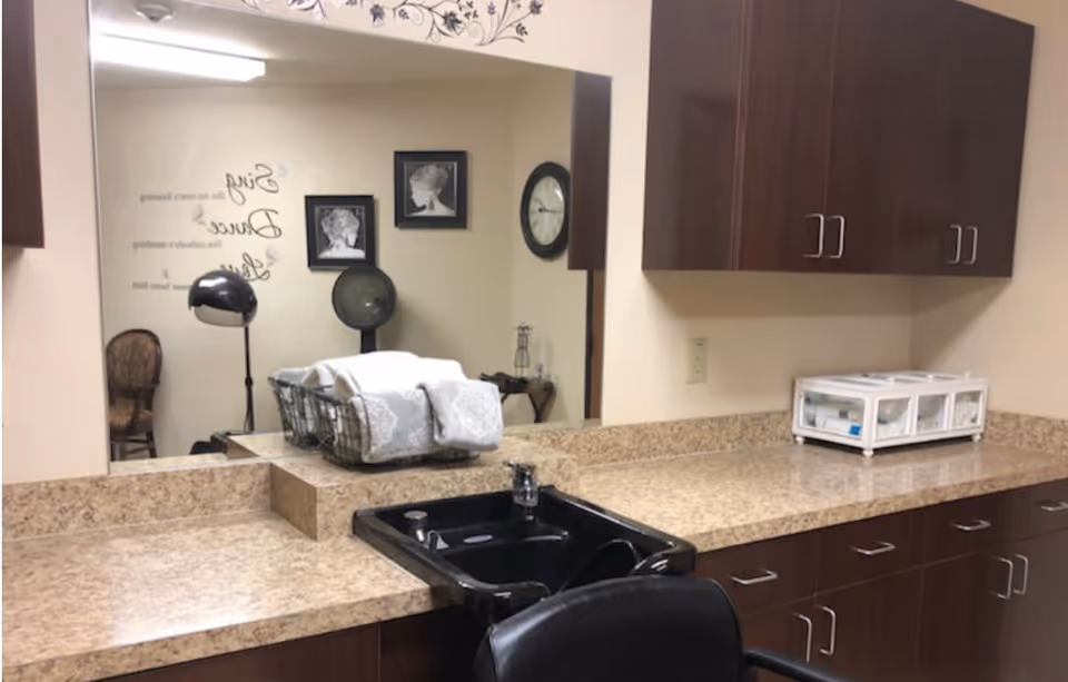 Interior view of a salon area in a healthcare facility featuring a black salon sink and chair, granite countertops, dark wood cabinets, a large mirror reflecting wall art and a hair dryer, and a basket with folded towels on the counter.