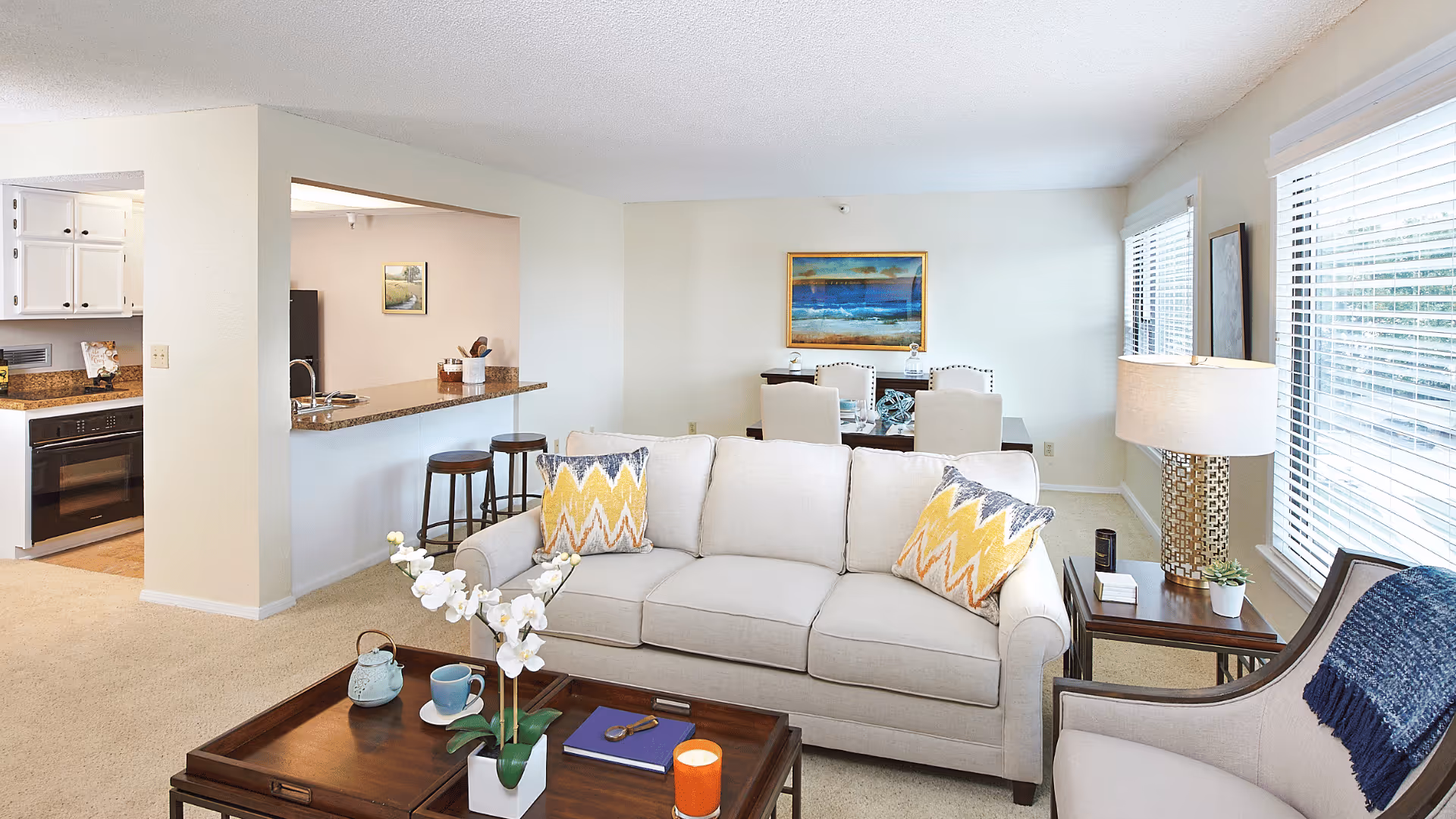 Bright and airy living room with a beige sofa adorned with yellow and gray patterned pillows, a wooden coffee table with a white orchid plant, a teapot, a cup, and a candle. To the right is a side table with a lamp and a small plant, next to a window with white blinds. In the background, there is a dining area with a table and chairs, and to the left, a kitchen with white cabinets and granite countertops.