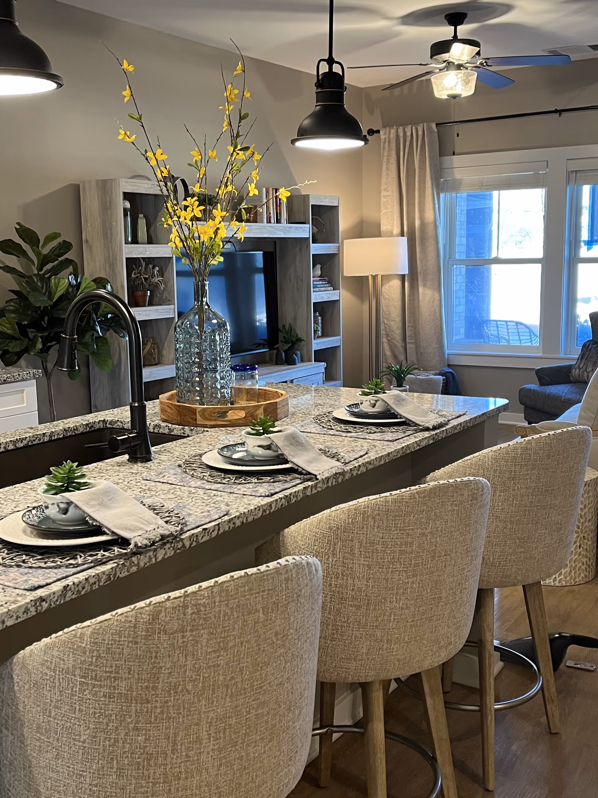 Interior view of a modern living space featuring a kitchen island with four beige upholstered bar stools. The granite countertop is set with plates, bowls, napkins, and small potted plants. A large glass vase with yellow flowers sits on a wooden tray in the center of the island. In the background, there is a living room area with a TV on a wooden entertainment unit, a floor lamp, a ceiling fan, and windows with curtains letting in natural light.