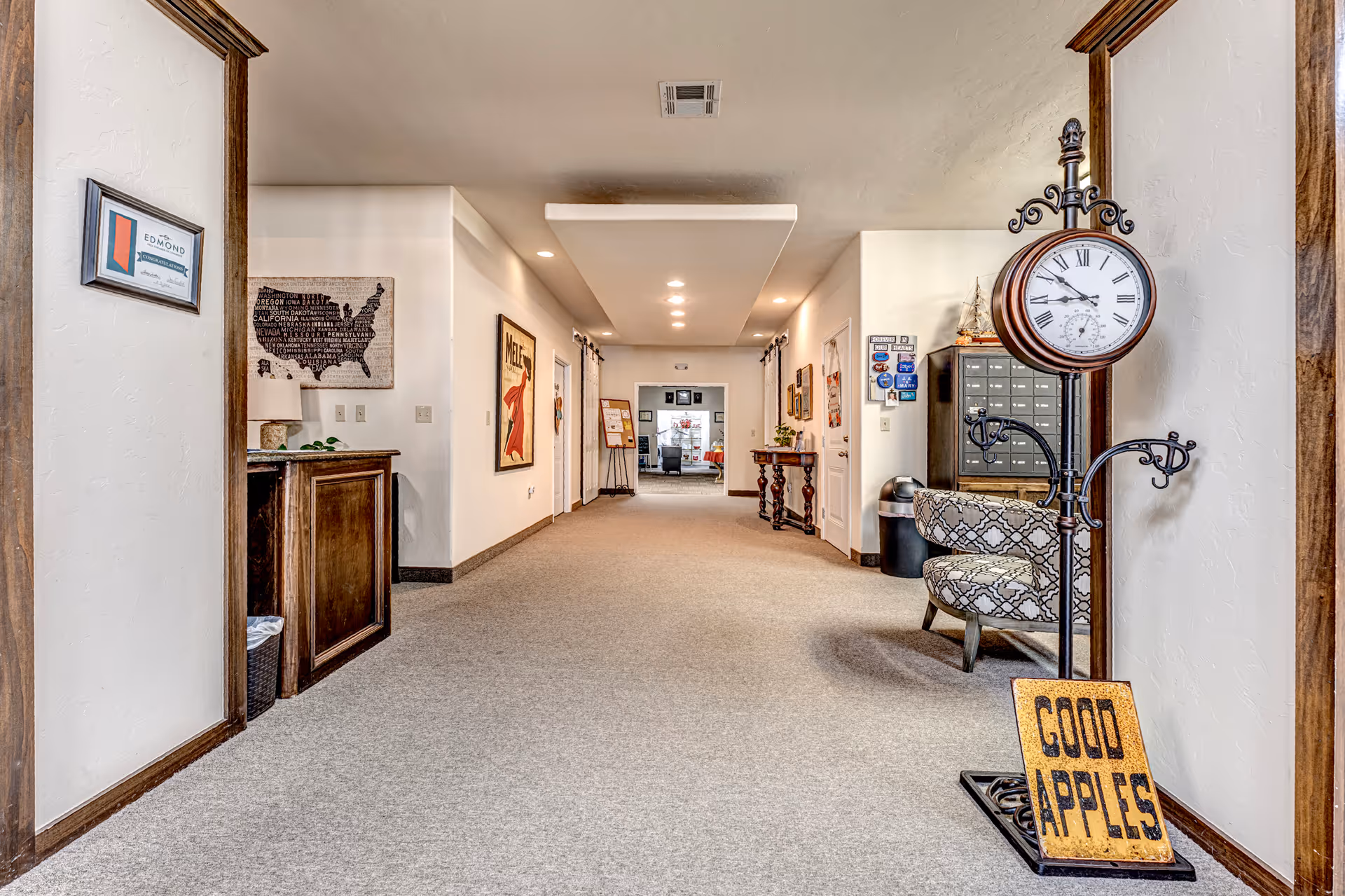 A carpeted hallway in a senior living facility with white walls and wooden trim. The hallway features framed artwork on the walls, a decorative standing clock with a sign that reads 'GOOD APPLES,' a patterned chair, and a small wooden table. The ceiling has recessed lighting, and the hallway leads to another room in the distance.