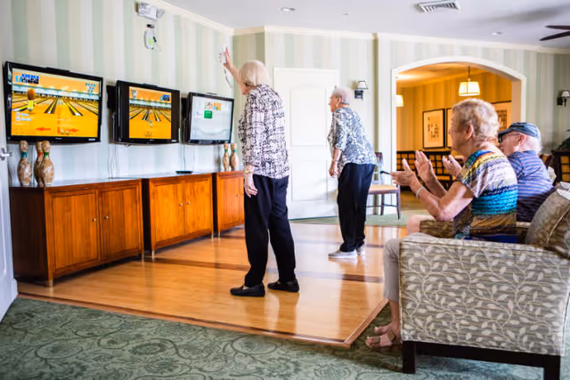 A group of elderly people in a common area of a senior living facility playing a virtual bowling game on three wall-mounted screens. Two women are standing and actively playing, while two others are seated on a patterned couch, watching and clapping. The room has wooden flooring, green carpet, and light-colored striped wallpaper.