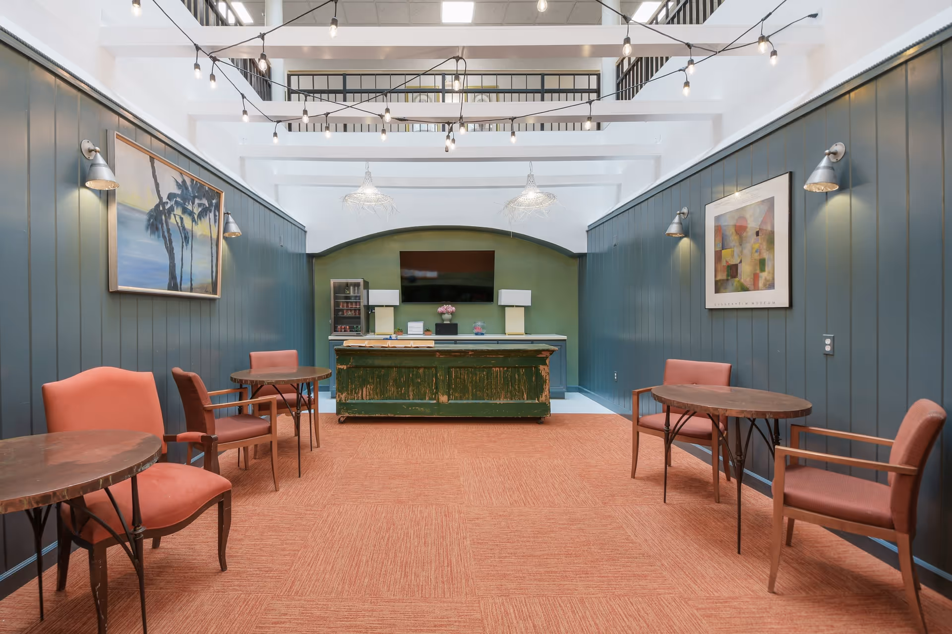 A cozy common area with orange carpet and dark green paneled walls. The room features several small round tables with orange upholstered chairs. On the far wall, there is a rustic green counter with two lamps, a small refrigerator, and a flat-screen TV mounted above. Two framed artworks hang on the side walls, and string lights are suspended from white ceiling beams.