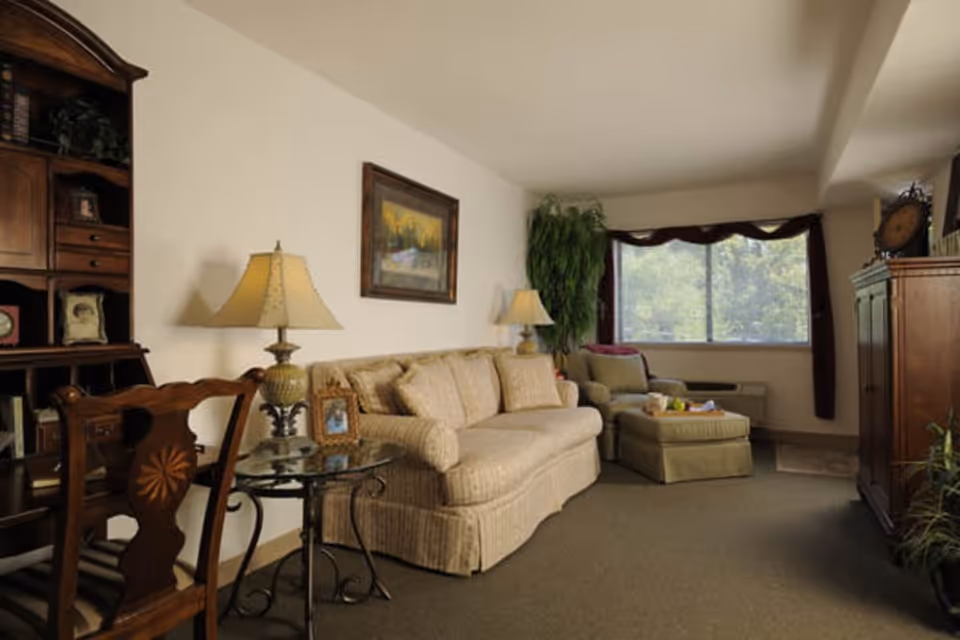 A cozy living room in a senior living facility featuring a beige sofa with matching cushions, a glass side table with a lamp and framed photo, a wooden chair, a large window with dark curtains, a green armchair with an ottoman, a tall green plant, and wooden furniture including a cabinet and a bookshelf.