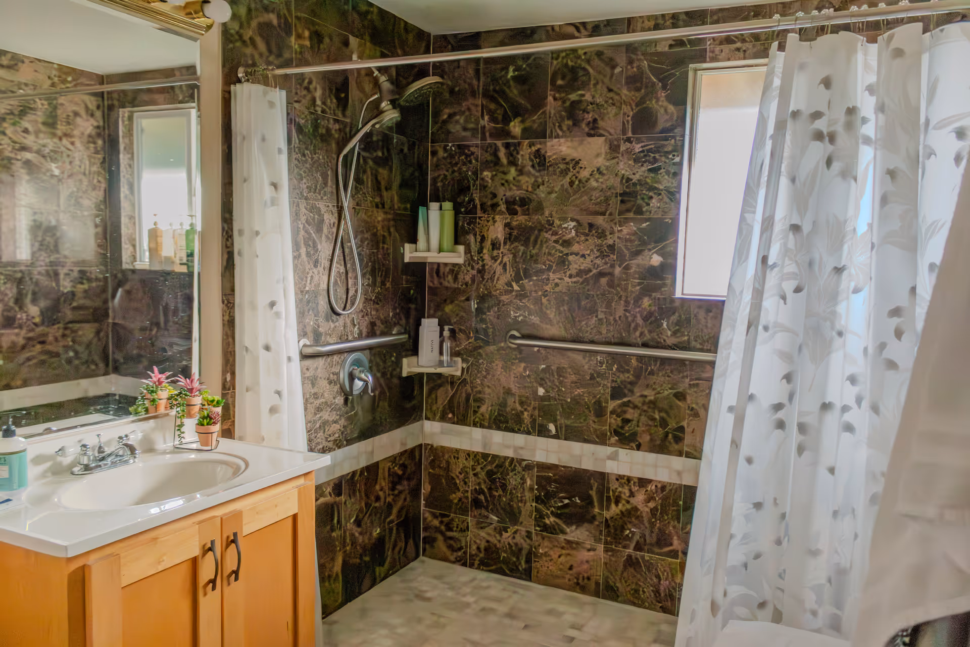 A bathroom with dark marble-patterned tiled walls, a shower area with a handheld showerhead, grab bars, and a white shower curtain with a floral pattern. There is a wooden vanity with a white countertop and sink, a large mirror above the sink, and small potted plants on the countertop. A window allows natural light into the room.