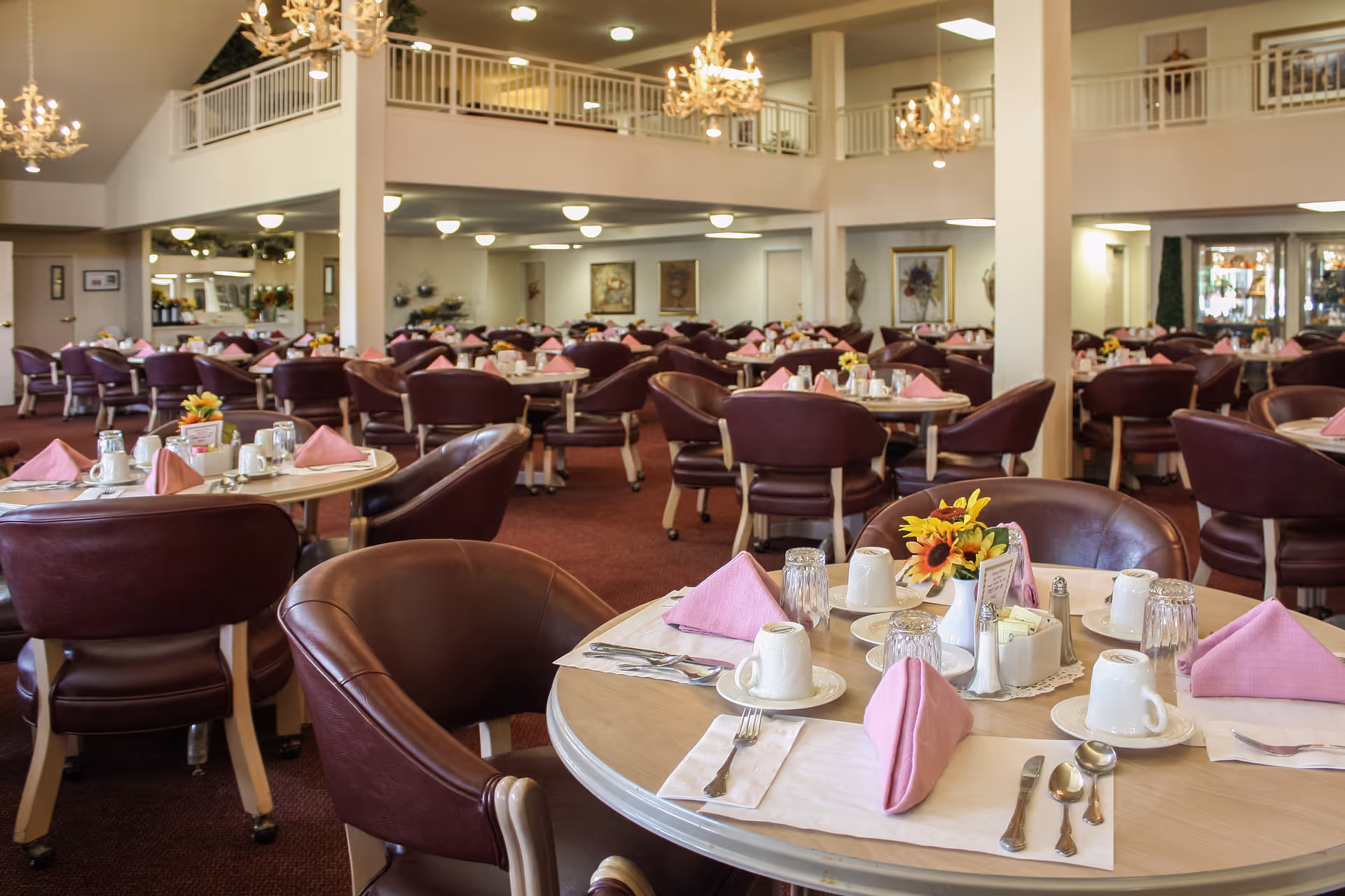 A spacious dining room with multiple round tables set for a meal. Each table has pink folded napkins, white cups, glasses, silverware, and small flower arrangements. The room features brown leather chairs, chandeliers hanging from the ceiling, and a second-floor balcony overlooking the area.