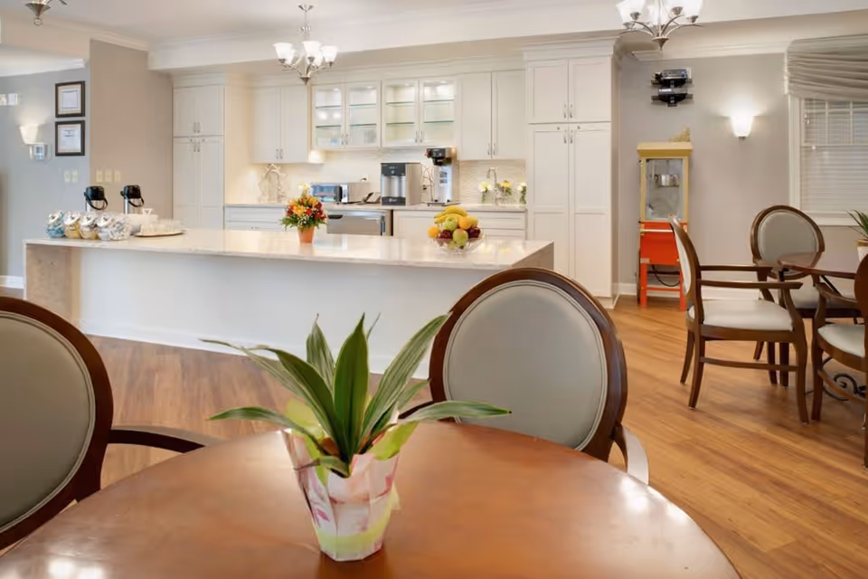 A bright and clean kitchen area with white cabinets and a large island countertop. The island has a fruit bowl and a flower pot on it. In the foreground, there is a round wooden table with a small potted plant. Several chairs with light-colored cushions surround the tables. The floor is wooden, and there is a popcorn machine in the corner near the wall.