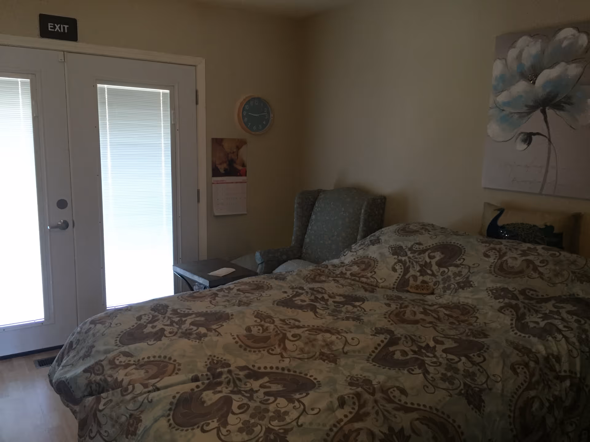 A bedroom with a bed covered in a patterned comforter, a cushioned armchair next to a small table, a wall clock, a calendar, and a large floral painting on the wall. There are double doors with blinds and an exit sign above them.