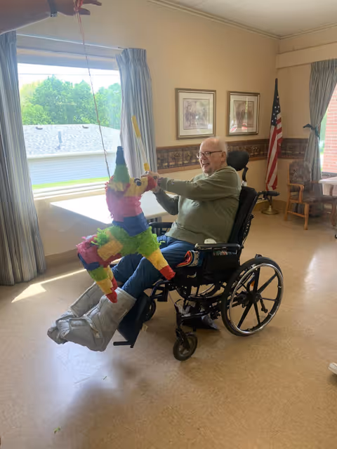 An elderly man in a wheelchair is smiling and holding a colorful piñata shaped like a donkey inside a well-lit room with large windows, framed pictures on the wall, an American flag, and chairs in the background.