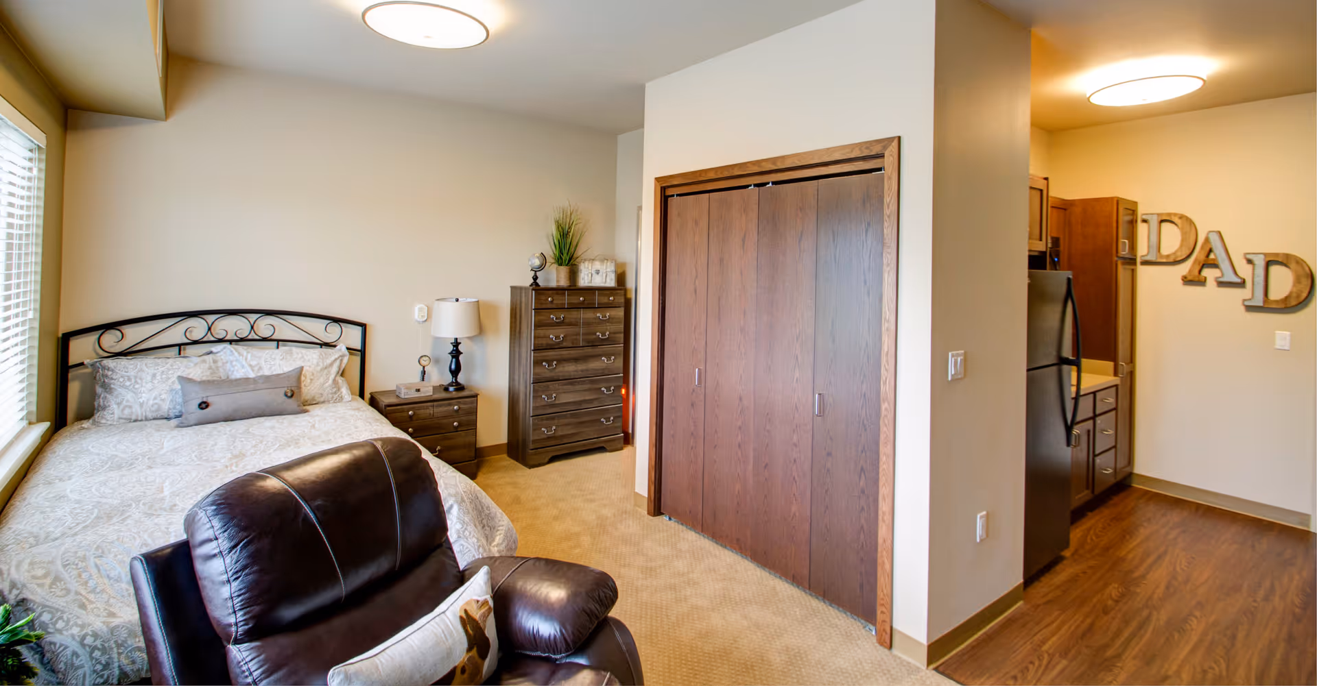 A cozy bedroom with a bed featuring a decorative metal headboard, a nightstand with a lamp, a tall dresser with decor items, and a leather recliner chair. Adjacent to the bedroom is a small kitchen area with wooden cabinets, a refrigerator, and wall decor spelling 'DAD'.