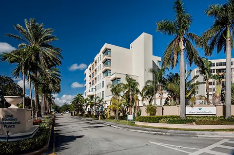 Exterior view of a multi-story building surrounded by palm trees under a clear blue sky. The building is part of Miami Jewish Health, located at One Egmont Sonderling Place, with a road leading up to the entrance.
