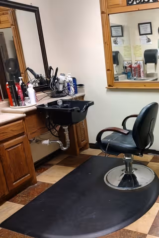 Interior view of a hair salon station with a black salon chair on a black mat, a black hair washing sink attached to a wooden counter, various hair care products on the counter, and a large mirror on the wall reflecting part of the room.