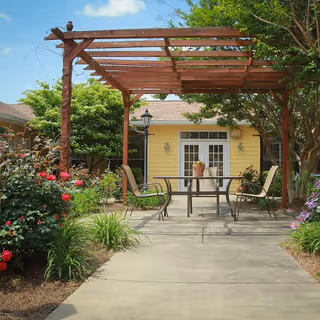 Outdoor patio area with a wooden pergola overhead, a round glass table with four chairs, surrounded by green bushes and flowering plants, leading to a yellow building with white double doors and windows.