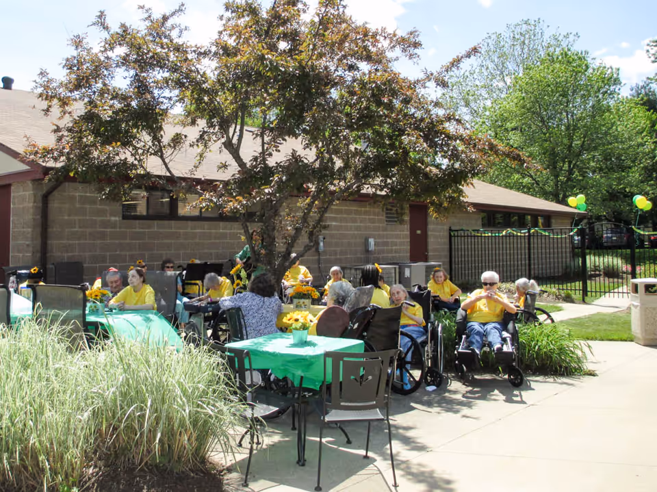 A group of elderly people, many in wheelchairs, sitting outdoors around tables covered with green tablecloths and decorated with yellow flowers. They are gathered under a tree near a building with a brick exterior. Some people are wearing yellow shirts, and there are green and yellow balloons tied to a fence in the background.
