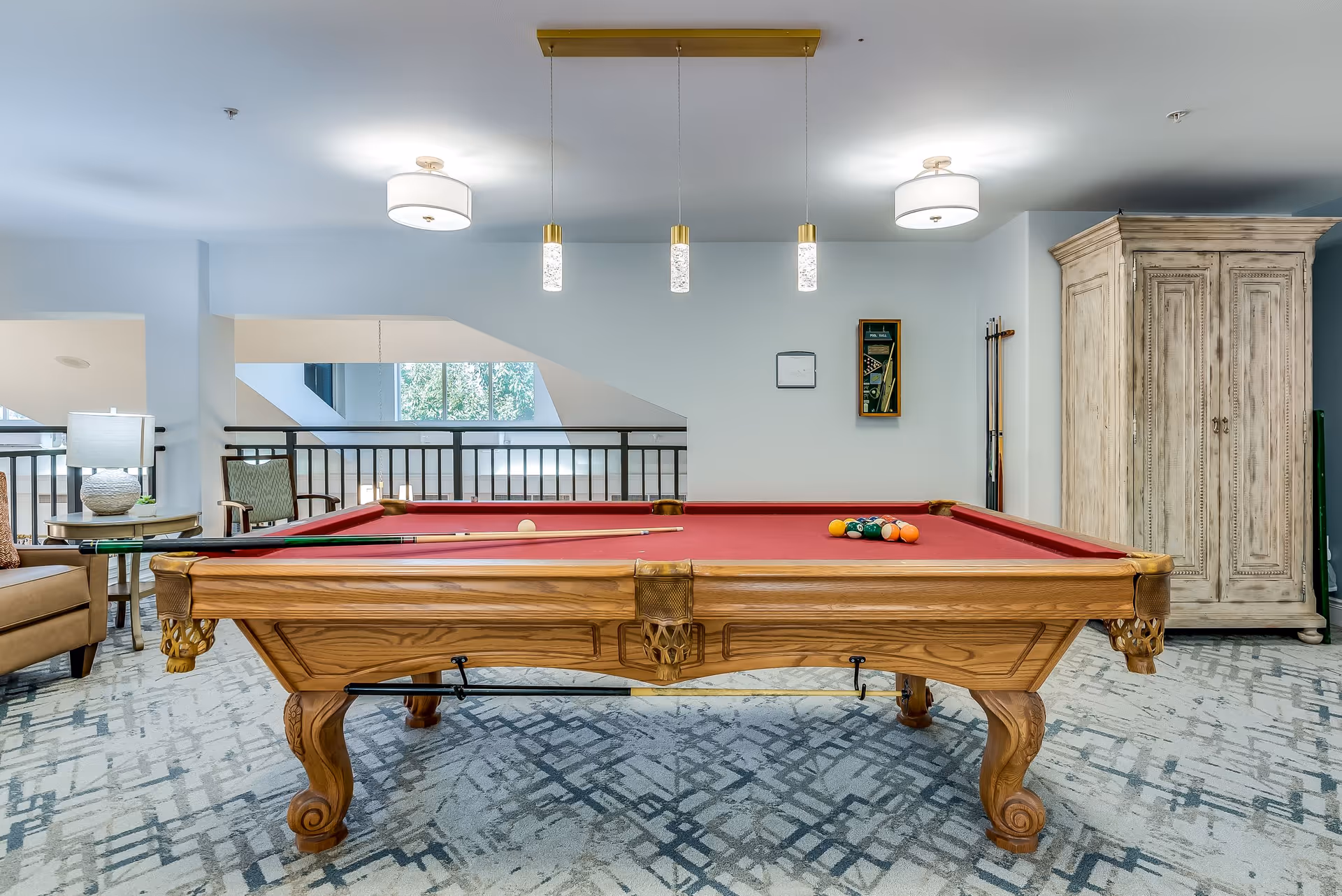 A well-lit room featuring a wooden pool table with a red felt surface, pool cues, and billiard balls arranged on the table. The room has patterned carpet flooring, a large wooden cabinet, a wall-mounted rack holding pool cues, and modern ceiling lights. There is a seating area with chairs and a side table with a lamp visible to the left.