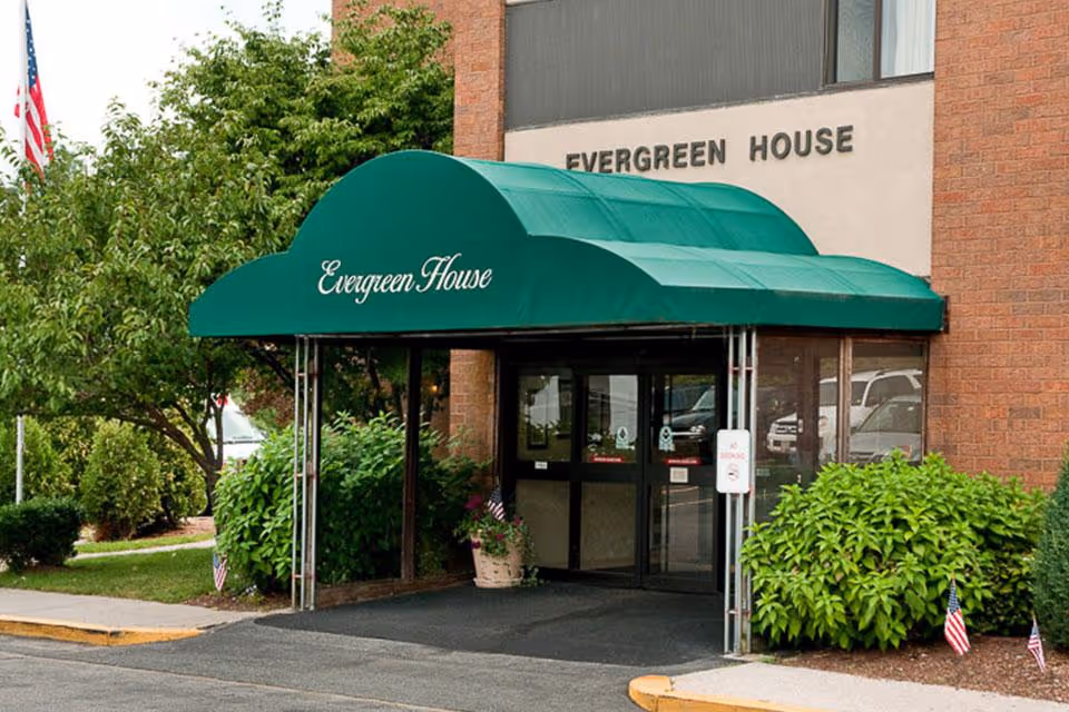 Entrance to Evergreen House Health Center with a green awning displaying the name 'Evergreen House'. The building is made of brick and has glass doors. There are bushes and small American flags near the entrance.