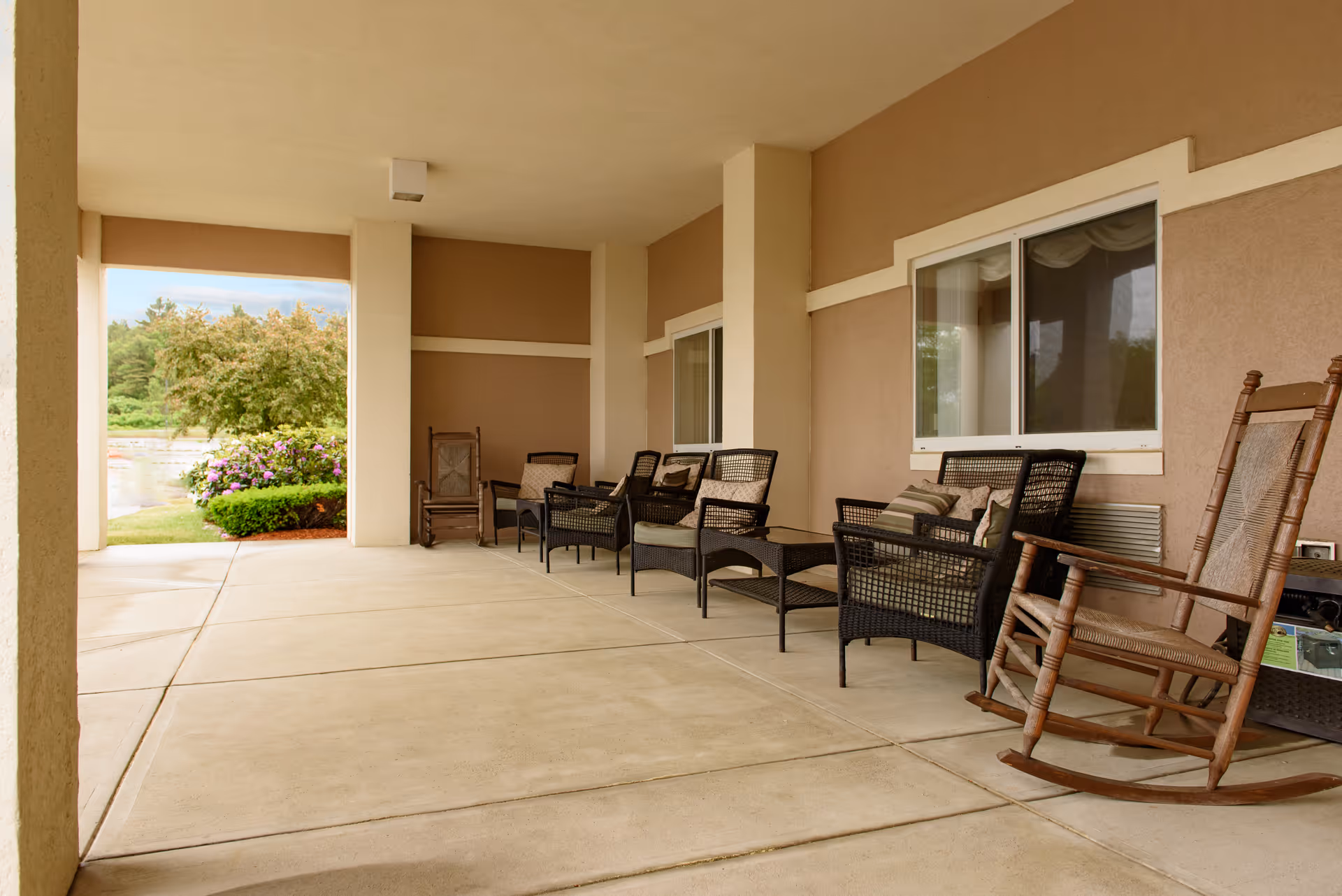 Covered outdoor patio area with several wicker chairs and wooden rocking chairs arranged along the wall. There is a view of greenery and flowering bushes outside the patio.