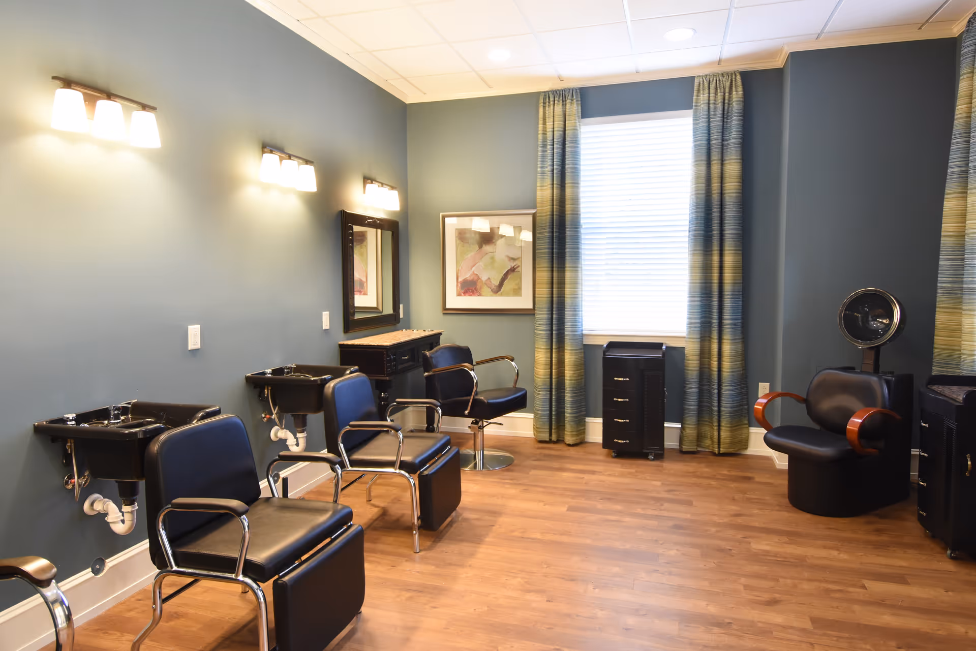 A bright hair salon room with styling chairs, wash sinks, mirrors, a hood dryer, and large window with curtains.