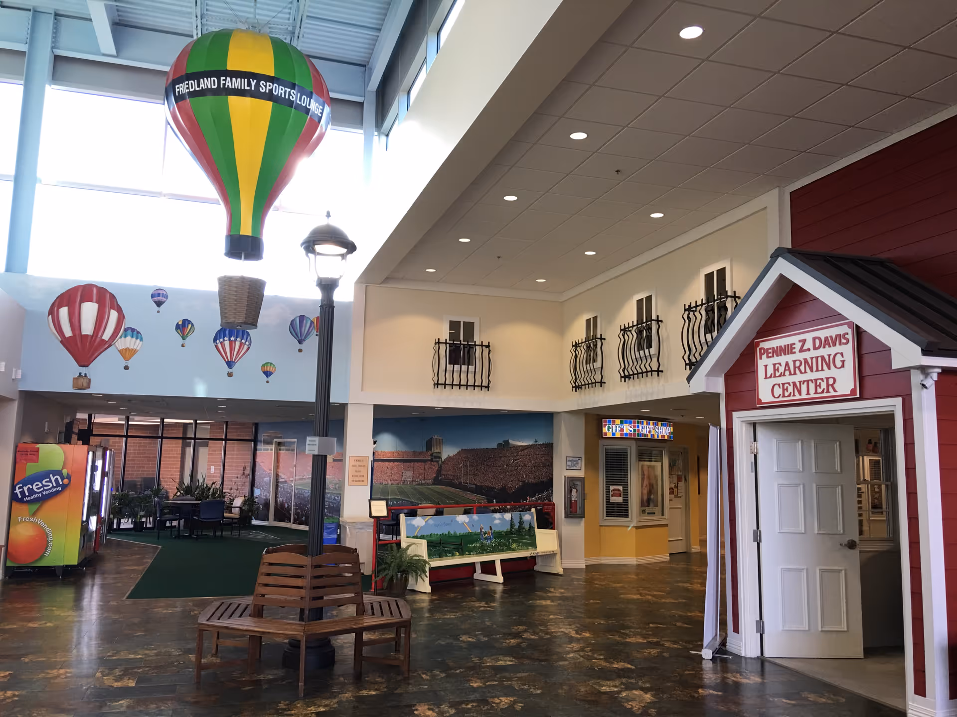 Spacious interior common area with hanging hot-air-balloon decorations, a small red building labeled 'Pennie Z. Davis Learning Center', benches, a vending machine and seating areas.