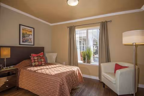 Cozy bedroom in a senior living facility with bed, chair, nightstand, and window.