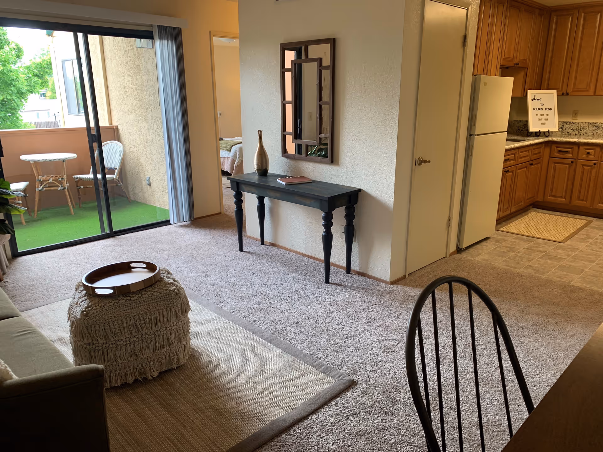 Interior view of a retirement community apartment showing a living area with a sofa, a textured ottoman with a wooden tray, a black console table with a decorative vase and a book, and a wall mirror. To the right is a kitchen with wooden cabinets, a refrigerator, and granite countertops. A sliding glass door leads to a small balcony with two wicker chairs and a round table, overlooking greenery outside.