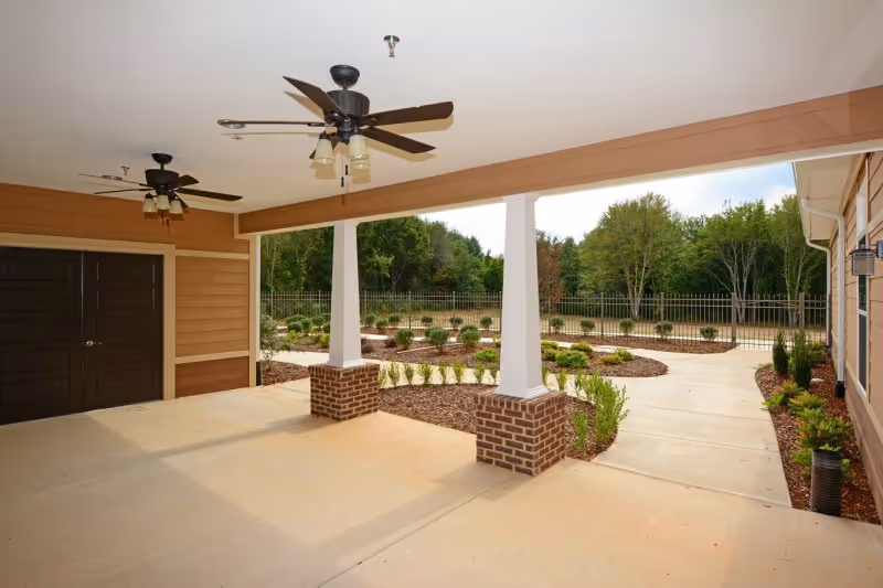 View of the entrance patio at Webb House Retirement Center, featuring ceiling fans, brick columns, and landscaped garden area with walking paths.