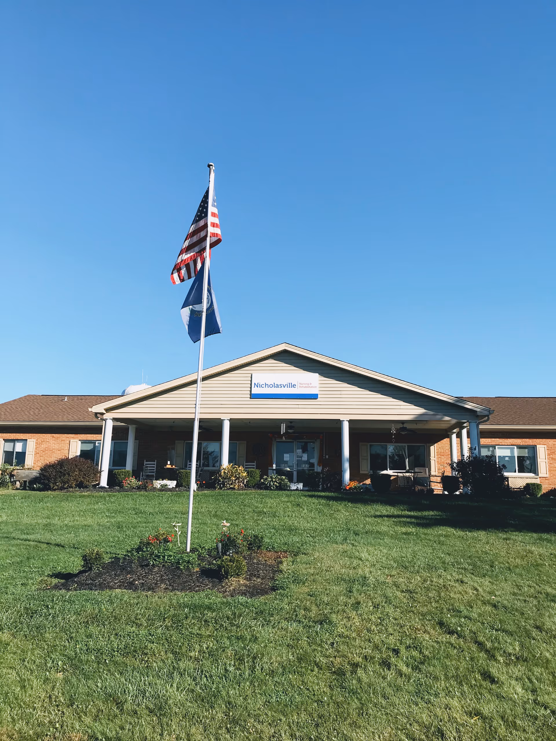Front exterior view of Nicholasville Nursing and Rehabilitation building with a well-maintained lawn and two flagpoles displaying the American flag and another flag under a clear blue sky.